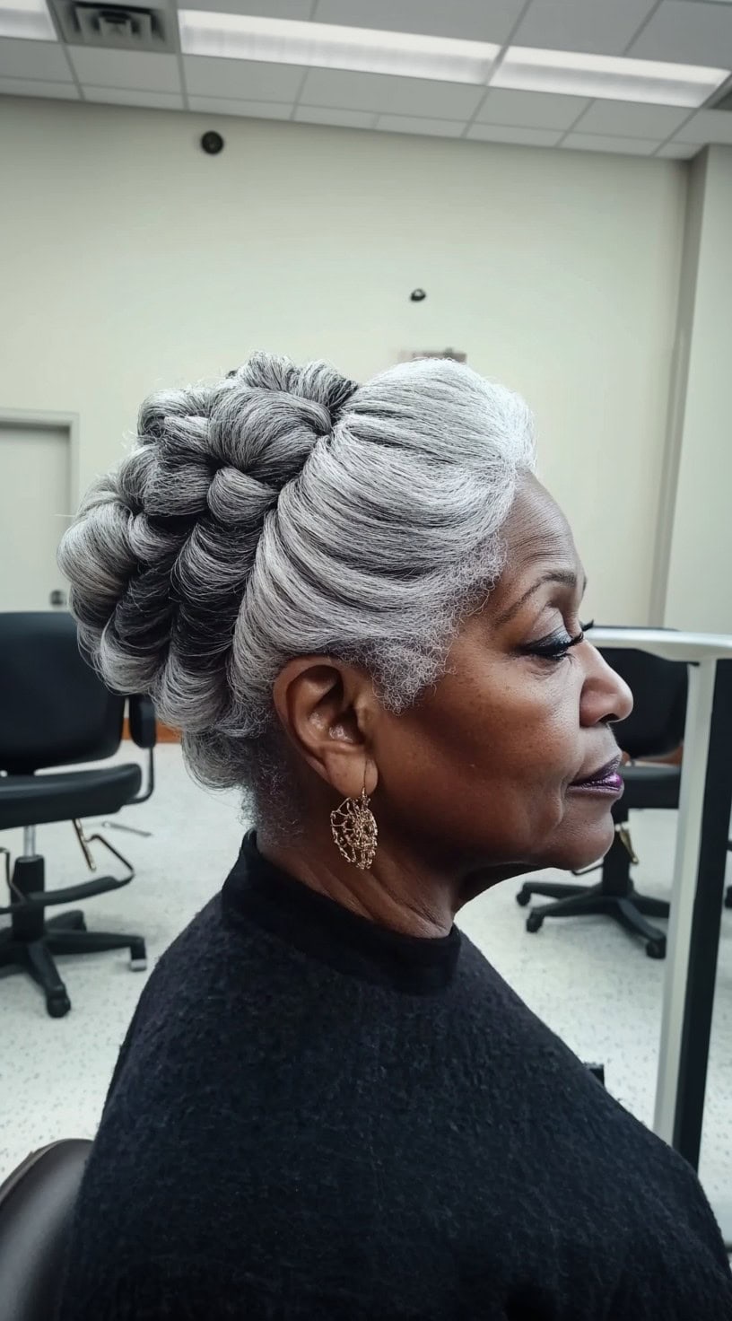 A side-profile view of an older Black woman with silver hair styled into an intricate braided crown updo, secured at the back.